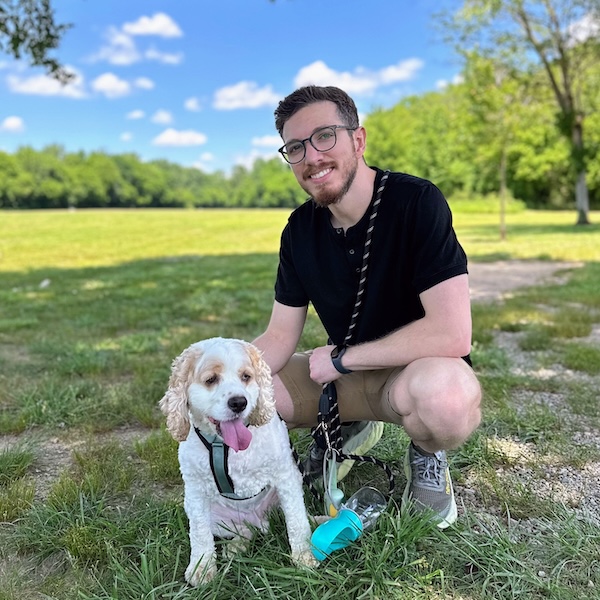 Aldo crouches on grass in a sunny park next to his dog, Bruno, a white and tan cocker spaniel with curly fur and his tongue out. Bruno is on a leash, and the background shows trees, a blue sky with scattered clouds, and a wide open field.